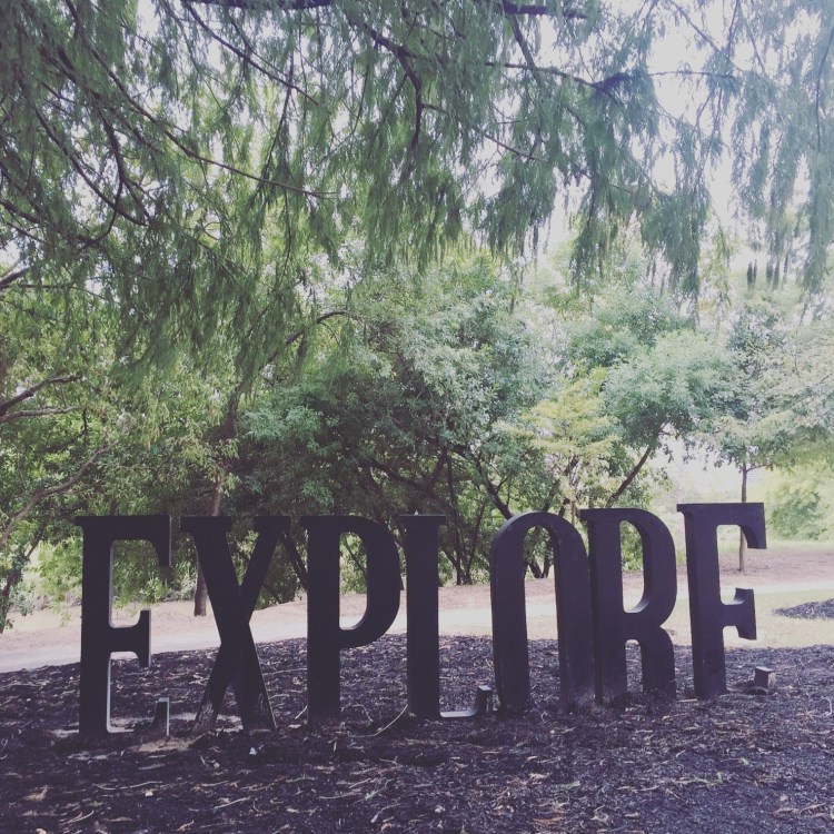 Photo is of a sculpture that says EXPLORE in capital letters. Green trees are in the background.