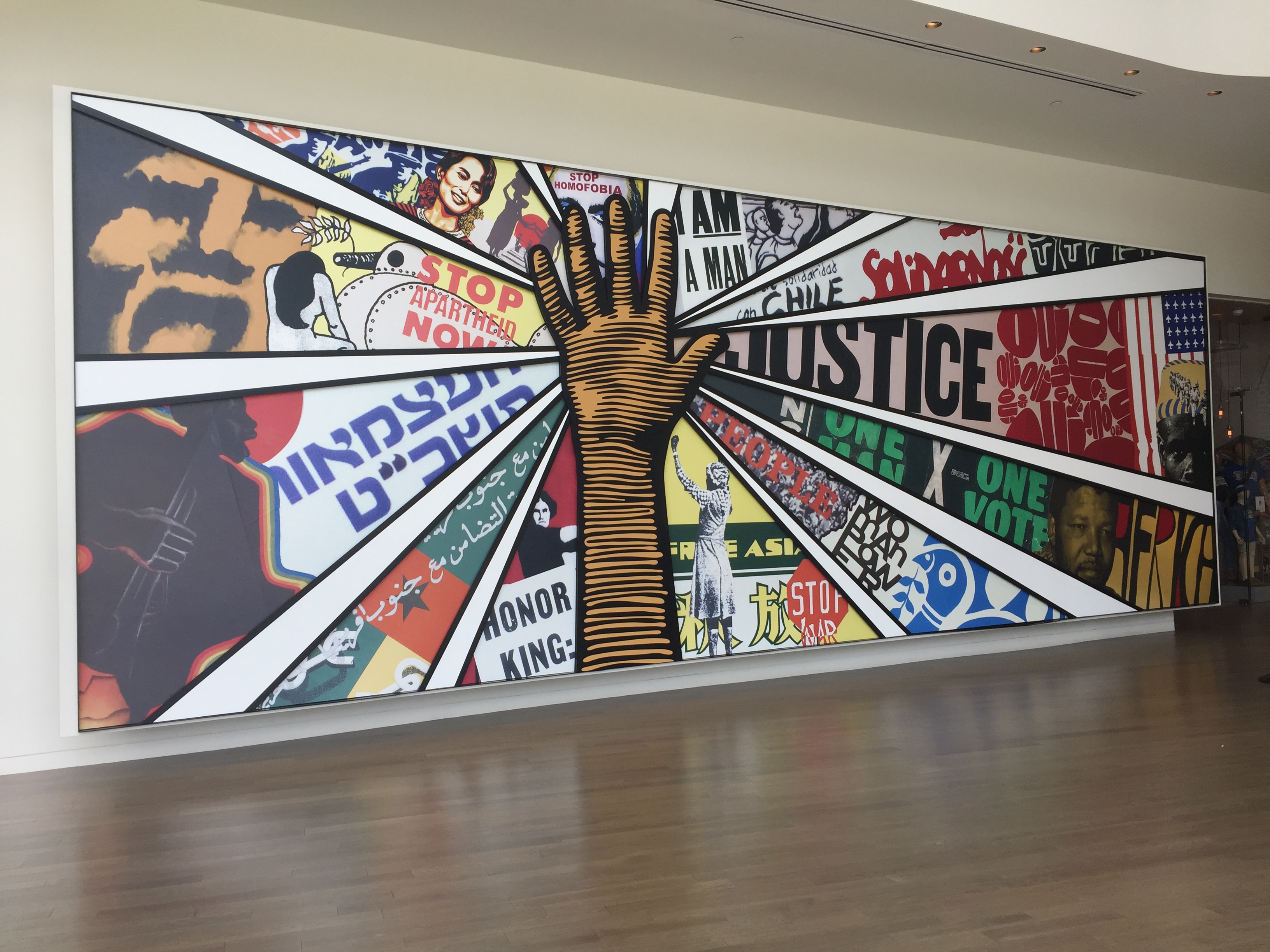 Photo is of a painting that has a hand in the middle and segments of colorful images meeting that hand in the middle. One of the segments says "Justice." Another says "Stop Apartheid Now."