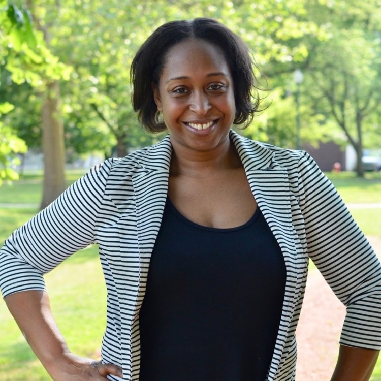 Photo is of Melody from the waist up. She is standing and wearing a black shirt and a black-and-white striped blazer. Her hands are on her hips. Trees are in the background.